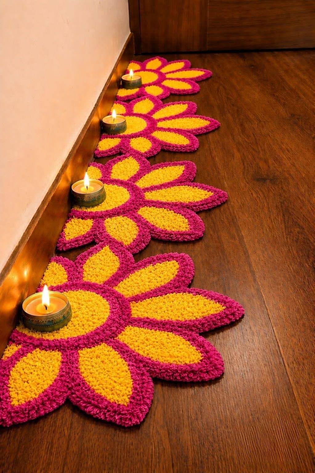 Four magenta and mango-yellow flower rangoli mats laid along a wooden floor with a lit tealight at each centre.